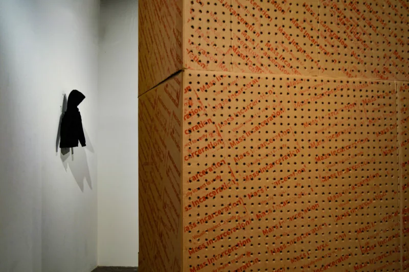 A photo of two art pieces in a gallery shows a small black hoodie mounted on the left wall, dwarfed by two large cardboard boxes stacked high and towering over the small hoodie. The boxes are printed with the words “Mass Incarceration” repeated diagonally on each side of the box.