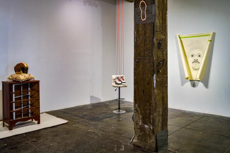 A group of four art works sit in the corner of a gallery, in which a rusted metal pillar occupies the foreground.The art includes two found-art pieces, one with white Air Jordan sneakers, one with a football helmet and one that is a drawing on a curled piece of paper or canvas of a Black man with a mustache. The fourth is an outline in pink tape of a foot placed high on the metal pillar.