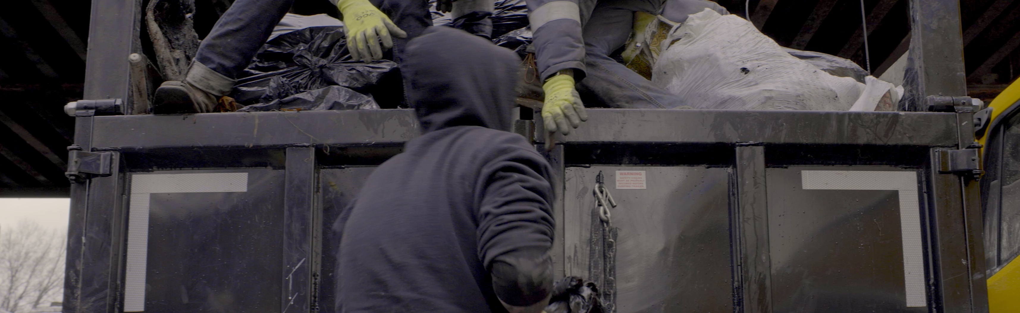 In a film still from Melissa Langer's film, "In Excess," a dark photo shows partial views of two men — one with his back to you and wearing a dark hoodie, and the other seen only with a foot and part of a leg showing and hands in yellow worker gloves either reaching down to receive something from the hooded man or to give them something. They are working with what looks like trash in a dumpster. 