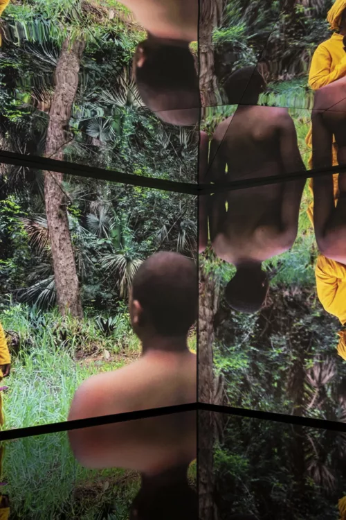 A photo shows a still image of a video on two wall size screens and reflected in a series of mirrors on the ceiling, the still image shows two screens featuring a young black man from the back who is shirtless, he looks towards a lush forest of green with a person dressed in yellow with a matching turban to his left, this image is reflected in the mirrored ceiling.