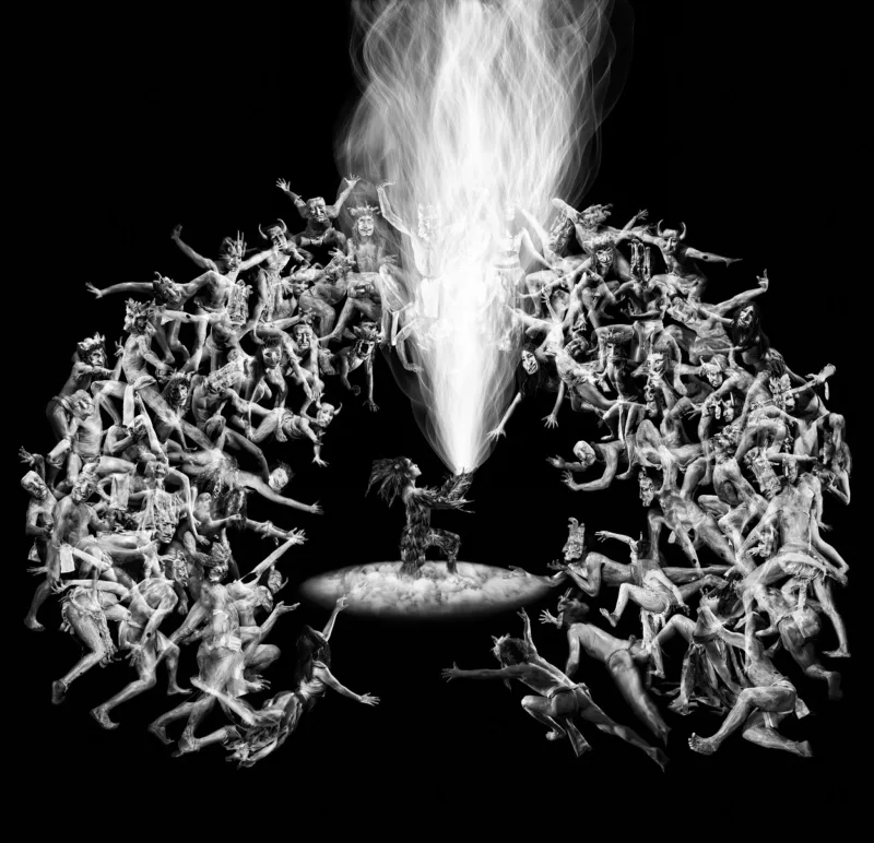A square black and white image. At the center is an almond shaped elliptical with a person in feathered dress shoots fire like spark trails from an object in their hands. Around the fire the formation of a house shoe of people in variety of poses and costume surround.