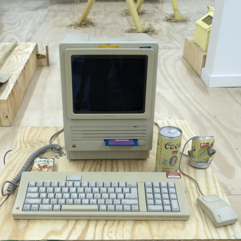 An old Macintosh computer with keyboard and mouse sit on a plywood table. In the space is a crushed Coors can alongside an unopened one. A pack of Camel Blue cigarettes sits on the left side of the computer. In the background parts of other sculptures appear.