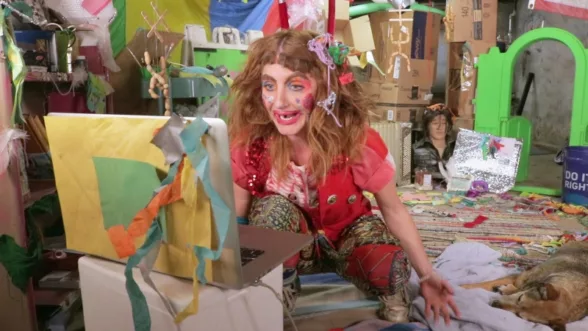 Artists Jenny Drumgoole as her persona Soxx looking at a laptop covered in colored paper sitting on top an ice chest. Soxx is dressed in red and patterns with gaudy makeup and large clips in their hair. The surrounding room is chaotic and filled with objects of household utility and colorful toys. A dog lays to Soxx's left.