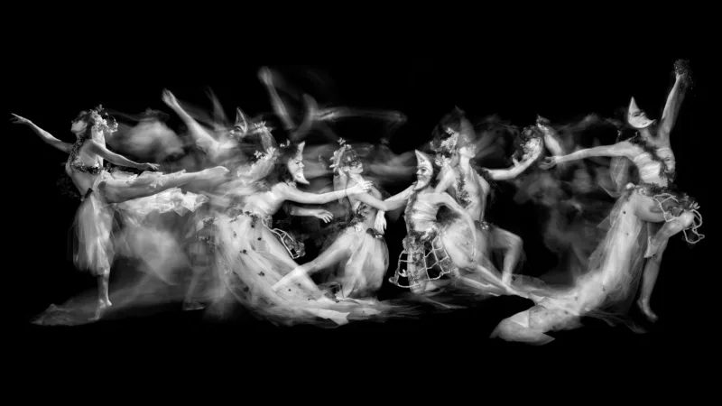 A horizontal canvas shows dancers in masquerade masks of humans and moons dance about. At the middle and right side the dancers are more still while moving lefts the motion of the dancers increases. At the center a female moon faced dance reaches for a moon face person.