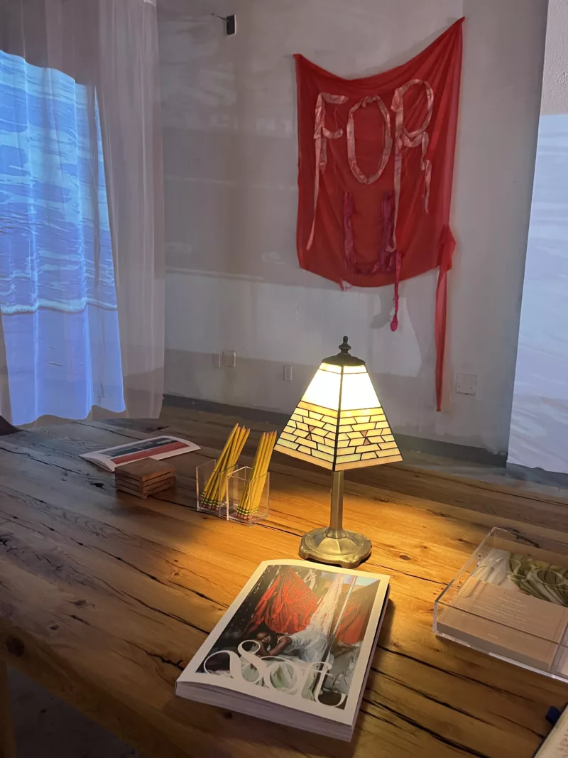 A reading nook set up on a wood table in the gallery space of the Asian Arts Initiative. On the table is the book Soft alongside a lamp, pencils, coasters, and some other miscellaneous items. In the background is the projection screen displaying a short film and a red banner hanging from the wall with the words "For U".