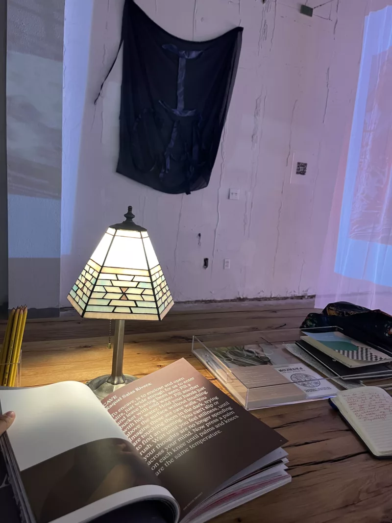 A reading nook in the Asian Arts Initiative gallery space. Set on a wood table with a glass lamp casting a warm yellow light over the new book Soft. In the background are wall hangings and a screen of translucent curtain which is being projected on.