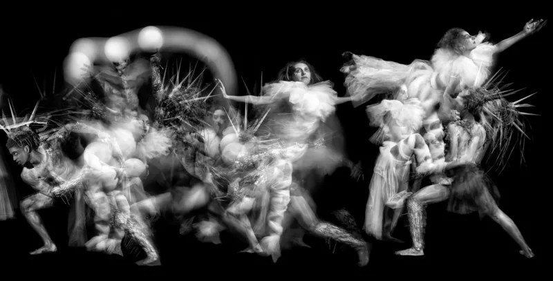 From left to right a horizontal stretch black and white photo. Dancers stretch from the bottom left towards the upper right of image, blurring and capturing the motion. A black man lunges towards the bottom left corner. To his right is a white blur of a dancer above a man with light orbs at their shoulders and head. In the middle the shadow of a woman rushes left and a woman leaps on point toward the right looking left. On the right a woman in a frilled costume is lifted into the air by a man with spiky hair.