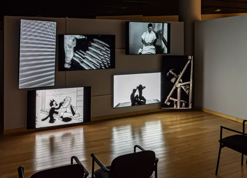 A picture of the gallery space with chairs place in front of six screens each displaying a video. On the upper left is an image of horizontals with grey in between the white slats, it almost looks like feathers. Below is a cartoon of a Felix the cat type character putting their finger in and old man's mouth. The middle left has an image of a man racking massive bullet shells. Below and on the right is a woman and man in crouch. Directly above is an image of a man looking at another person in a gurney. Directly right of the man and woman is an image of a mechanical device.