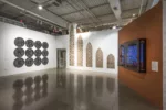 A photo shows a gallery room with artworks hung on opposite walls and one along another wall in the back. Each object has a repeating pattern. On the right is a window-like structure with two sashes in front of an image with vibrant blue background and a radiating pattern over it. this object hangs on a deep orange colored wall. Across from this object on the left is a series of 12 wooded circles with a grid pattern, each has letter that spells out WOMEN CAIRO 22. Along the back wall is a series of five of mashrabiya, or wood lattice structures that are square at the bottom and pointed at the top, these range in size from biggest on the left to smallest in the right; each has a similar radiating pattern within its frame.