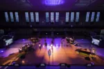 Students at Girard College rehearse Be Holding in the Armory gym at the school. Two pianos and drum kits flank the students in the middle. The floor and students are lit in orange and purple.
