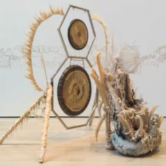 A large gong sits in an octagon metal frame with a hexagon and smaller gong on top. From the sides radiates some spiky yellowish white material. In front of the gongs is a collection of difficult to categorize objects sitting in a blackish white basket. Tendrils point out of the amalgamation. Behind on the gallery wall is a white/yellow series of squiggles overlapping.