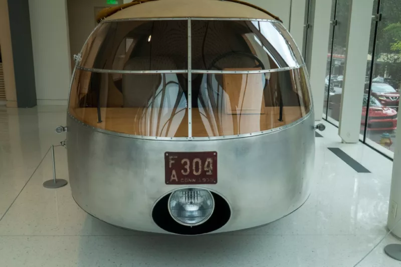 Front view of Buckminster Fuller's Dymaxion car. The car has four panel windows with a strip of metal riveting binding it together. The bottom is brushed metal with a large opening for a single headlight. The top is covered with a beige textile material. The interior shows two seats and a wheel with a light wood dash.