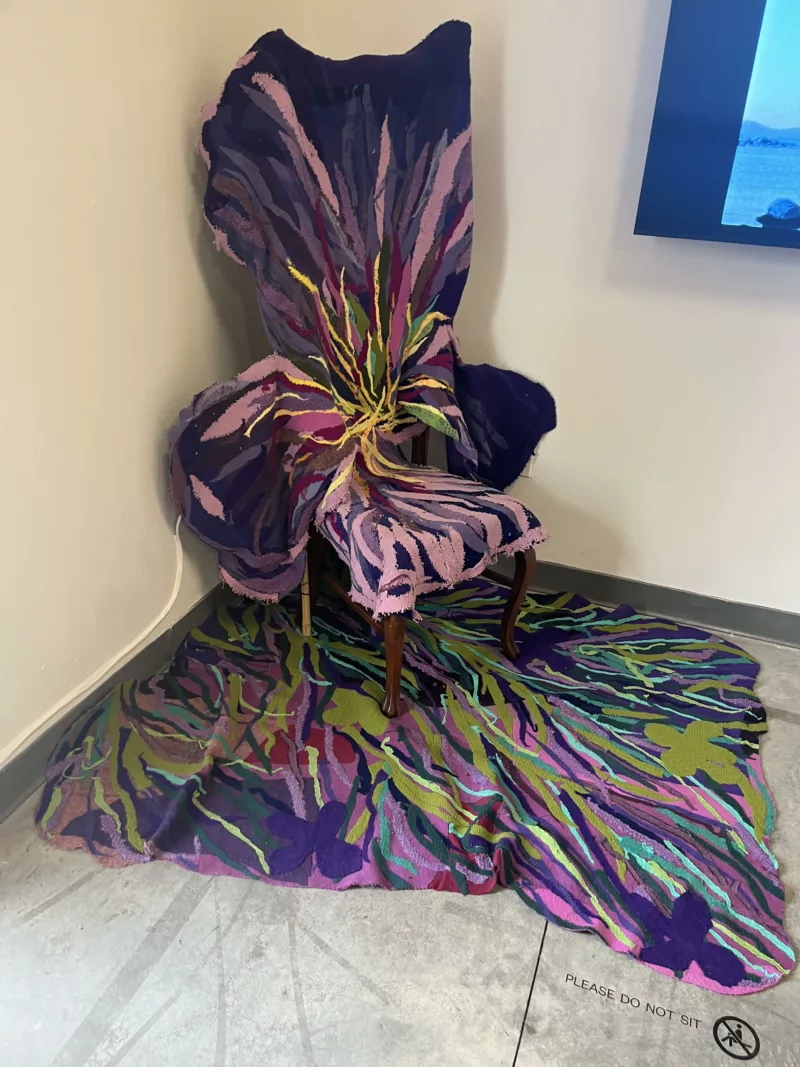 A high-backed chair sits in a corner, entirely covered with colored cloths and ribbons of fabric in beautiful colors seeming to emanate from the seat and spill over onto the floor covering a triangular area under the chair with the same colored fabric.