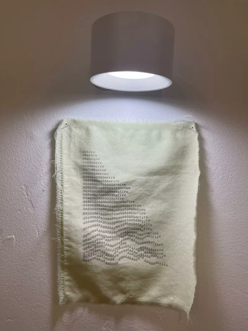 An off-white colored cloth hangs on a wall, spotlit by a large light. The cloth contains the words of a poem.