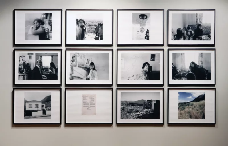 A grid of 12 images by Sue Williamson in 3 rows. 10/12 images are black and white. On the bottom row a piece of paper with writing and a colored image of a field and mountain appear. The top row of photos show a family together, the 3rd picture in the row shows a clock on wall and writing from the family. The middle row show's the individual family members in their home. The last row shows images of the home before and after destruction and a paper.