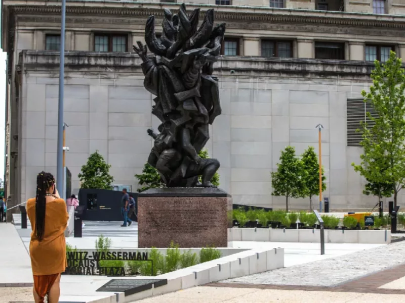 Image of the Horwitz-Wasserman Holocaust Memorial Plaza on 16th and Arch Streets. A woman stands in front taking a photo of the sculpture that sits in the plaza.