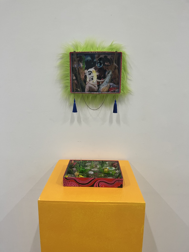 A gallery wall with a framed collage of people and statues with a furry green border, tassels hand off the bottom edges. Below is a yellow plinth with a little greenery landscape of plastic and found objects.