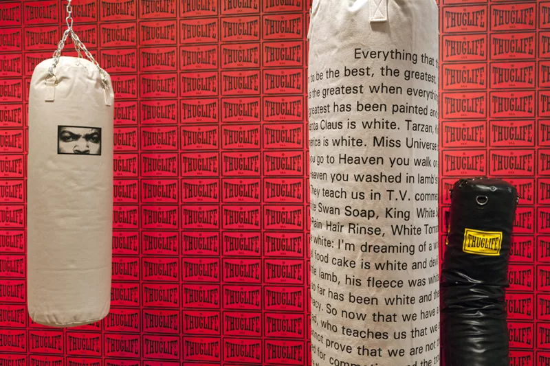 A work by Glenn Ligon titled "Skin Tight (Thuglife II)," it shows a room with a red rectangle with a flared text similar to TapOut saying "Thuglife". Punching bags hang from the ceiling and one on slumping in the corner. The left most bag is white with a rectangle cutout of a man's nose, eyes, and eyebrows. The man looks similar to rapper IceCube. The right bag has a scroll of text the right most bag is black and has a yellow thuglife sticker.
