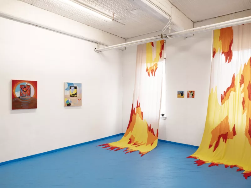 A corner of a gallery shows a bright blue floor with curtains draped from ceiling to floor in bright yellow and red.