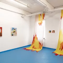 An image of the gallery during Seeing Red, it shows the painting along the wall. Red, orange, and yellow weavings with clear parts drip from the ceiling landing among the blue floor of the gallery.