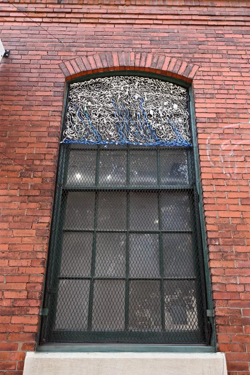 A window at the Crane Arts building with plastic wrapped electrical wires weaved into the grating of the window. The dominant color is white with a branched tree pattern of blue lining the bottom.