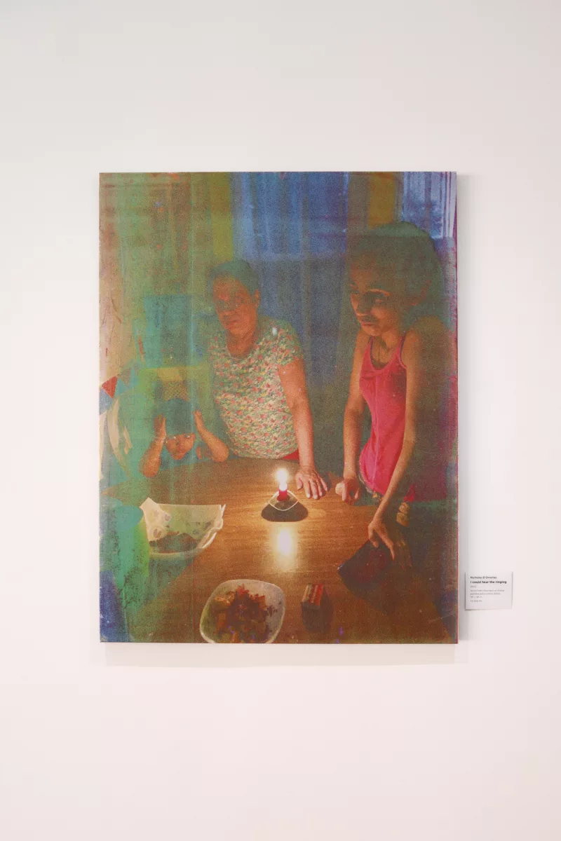 A woven fabric with silk screen print of an Indo-Caribbean family celebrating a birthday. The colors blue, magenta, and yellow stick out in color blocks. The skin of the people and the light of the candle illuminating them are also in vivid color. The image itself and it's weaved material gives a pointillist effect to the photo. On the left a little bold holds his head in his hands with his elbows on a round table. The table has a single pastry with candle in it and two other dishes of food. To the right of the boy is an older woman she rests here left hand on the table near the flame. A younger girl is on the right side of the frame in a magenta top, she looks off the edge of the frame.