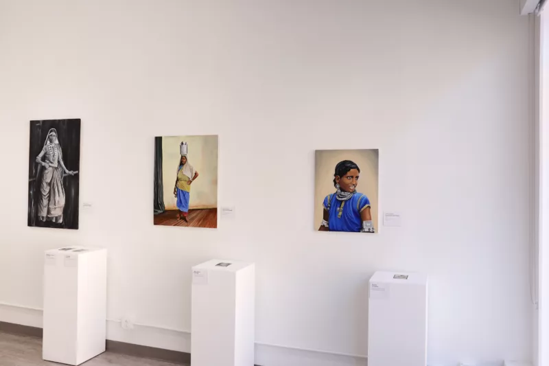 Three paintings of Indo-Caribbean women by Nazrina Rodjan. From right to left a portrait of a woman from chest up in blue clothes with silver jewelry adorning arms, neck, and ears the woman looks to her left. The middle painting shows a woman standing in a wood floored room, she stands with one hand on her hip and a jar atop her head. She wears a blue skirt with green top and beige head covering. To the very left a black and white of a woman in illustrious clothing with one hand on her hip and another on what appears to be a railing. Below each painting is an original image of the women depicted in the paintings, among the first laborers to arrive to the Caribbean from South Asia as laborers.