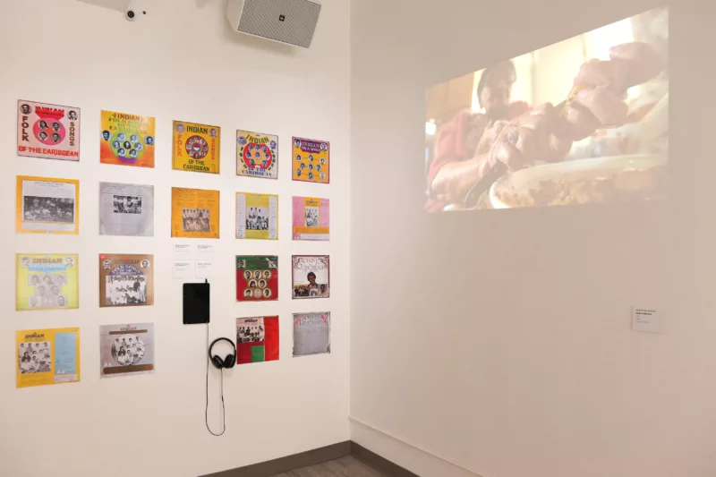 An image of two walls meeting to make a corner in the gallery space. On the left the curator installed Indo-Caribbean folk albums from their personal collection and a tablet with headphones for listening. To the right a short film titled Body Subpoena by Sarah Rohan Drepaul. In the shortfilm an older Indo-Caribbean woman cooks for a younger woman, and takes a look at the lives of these two people and the cultural artifacts and traditions that they engage with.