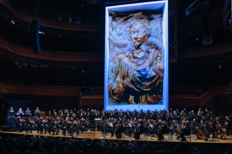A photo of the Philadelphia Orchestra playing beneath an AI generated image by artist Refik Anadol, the AI image shows a woman similar to that of Roman and Greek statuary but in an uncanny almost stitch like quality.