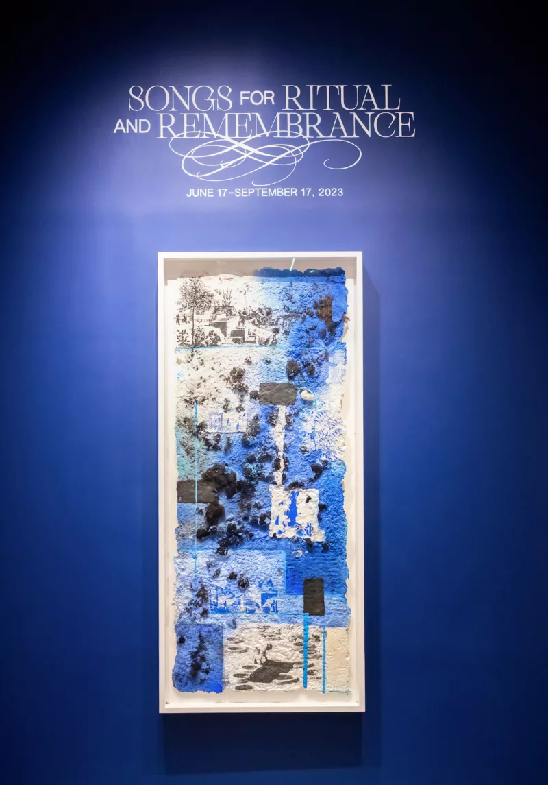 A dark blue wall announces an exhibit, with text in white at the top saying “Songs for Ritual and Remembrance” and, under the text, a framed work on paper echoes the blue of the wall with accents of white and squares of black.