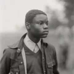 An image by Judith Joy Ross of a young black boy in a jean jacket with a sweater and collared shirt beneath. The boy stares off into the right side of the composition. The viewer sees both eyes and the features of his face in an almost profile pose. The background is blurred but there appears two men on the right side and a colonnade of trees.