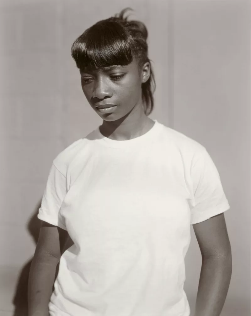 A black and white image of a black woman in a white t-shirt. The woman wears her hair back with bangs curtaining her forehead. She looks down toward the left corner of the border.