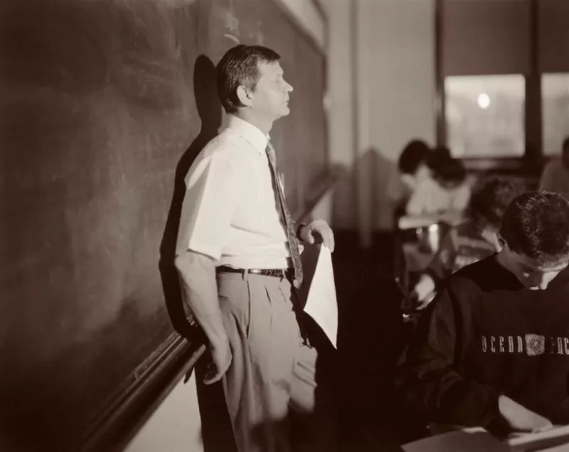 A black and white image of a white man with short dark hair. He is a teacher and leans on the chalk holder of his whiteboard. He looks horizontally across the composition over the heads of the students which intersect his eye line in perpendicular columns.