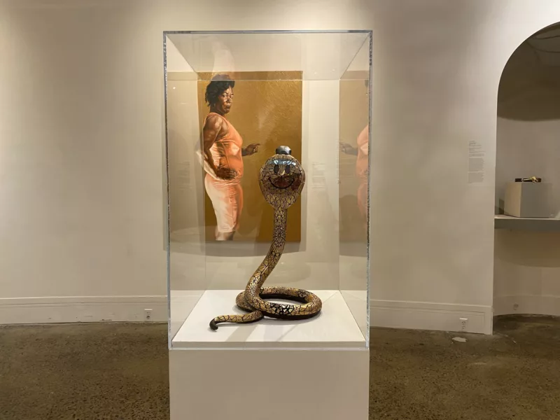 A gallery in Woodmere Art Museum shows two works, a painting of a Black woman in a pink dress, on a wall in the background and the rear of a 3D sculptural king cobra snake under glass on a pedestal.