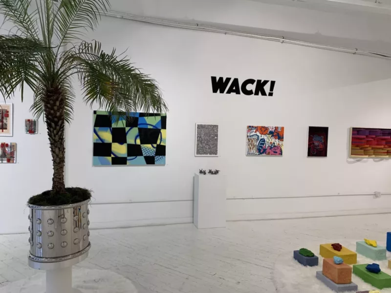 A photo of the exhibition WACK! at the HOT BED gallery space. The room is full of white walls with white wood flooring. To the left there's a palm tree in a silver planter with dots. On the wall are paintings, one with a plinth below holding small figures. The paintings vary in color, style, and even dimension. To the bottom right is a sculpture of colored objects.