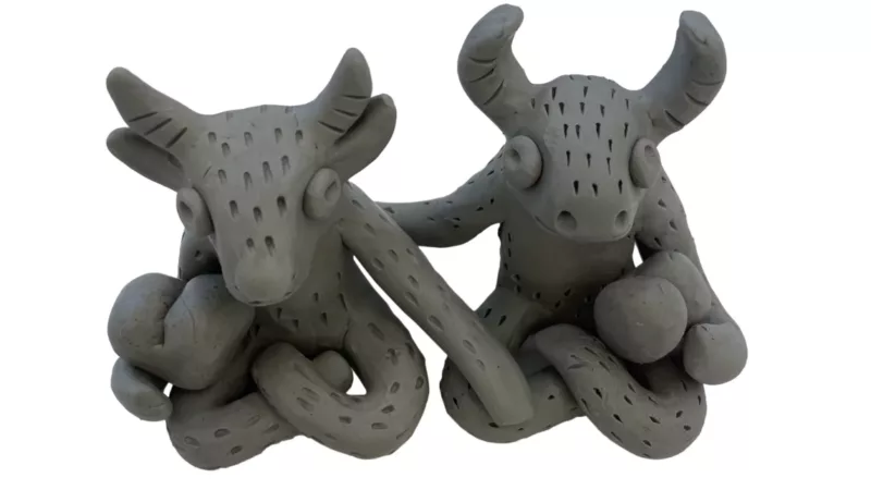 Two Roma Plastina modeling clay sculptures titled Little Guardians Nos. 96 & 97, by Aubrey Fink/Wolfdog. The sculptures are gray-green long eared, curved horned creatures that sit in lotus pose and touch one another with a hand as the other hand holds what looks like a pair of bongos.