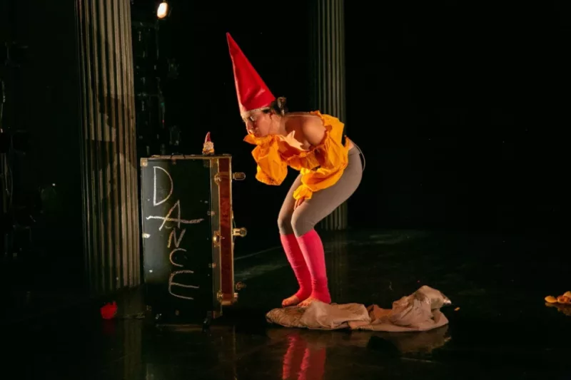 The artist Alexandra Tatarksy half crouches to a garden gnome on a stage box with hand drawn letters spelling Dance. Alexandra wears a cadmium yellow billowy top with grey leggings and pink leg warmers, atop their head is a pointed red gnome hat.