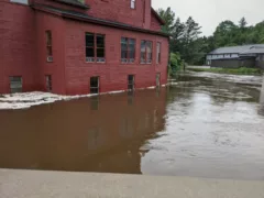 Vermont Studio Center buildings flooded. The image shows a red farm like building with windows, the water rising to the level of the windows, in the background two small buildings sit just above the massive pool of water.