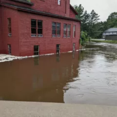 Vermont Studio Center buildings flooded. The image shows a red farm like building with windows, the water rising to the level of the windows, in the background two small buildings sit just above the massive pool of water.