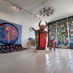 A gallery view of two tapestries and two figures. The largest figure is a horned upright figure with hot pink, yellow, and orange garb. It's long nails reach radiantly from its body. Below a smaller figure like that of a dinosaur stands with concentric colored circles overlapping and a pink face. To the left is a tapestry the drapes from the wall to the floor. The colors are vibrant with similar concentric circles as the figure. An oculus with hanging black tassels shows the vision of a mythic scene in great and vibrant colors. The tapestry on the right depicts a tree of life, creating a vibrant explosion of flora and fauna within it's foliage.