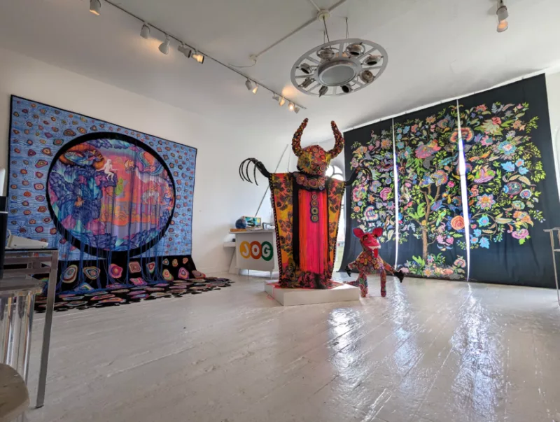 A gallery view of two tapestries and two figures. The largest figure is a horned upright figure with hot pink, yellow, and orange garb. It's long nails reach radiantly from its body. Below a smaller figure like that of a dinosaur stands with concentric colored circles overlapping and a pink face. To the left is a tapestry the drapes from the wall to the floor. The colors are vibrant with similar concentric circles as the figure. An oculus with hanging black tassels shows the vision of a mythic scene in great and vibrant colors. The tapestry on the right depicts a tree of life, creating a vibrant explosion of flora and fauna within it's foliage.
