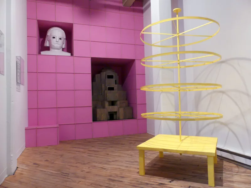 Photo of a installation in Grizzly Grizzly by David Herbert. It features a yellow table with a rod and wheels around, a pink wall of boxes, with two niches one holding a white bust another a wood temple like structure.