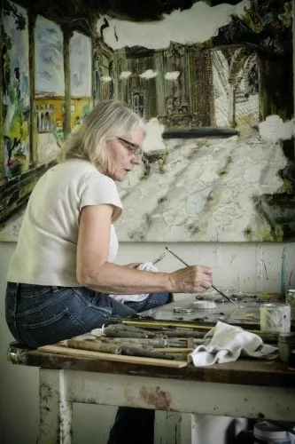 A grey haired woman sitting at the front of a palette and canvas.