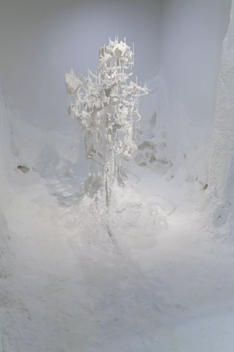 A white space with flocking bridging the space between the gallery floor and walls, creating a snowlike effect. From the center a delicate structure appears creating a cathedral of white.