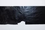 A large horizontal work on paper that is completely black and shiny sits on a white wall in a gallery. There is a jagged tear in the paper in the middle of the piece, which shows the effects of the artist working the materials very hard, resulting in tearing the paper.