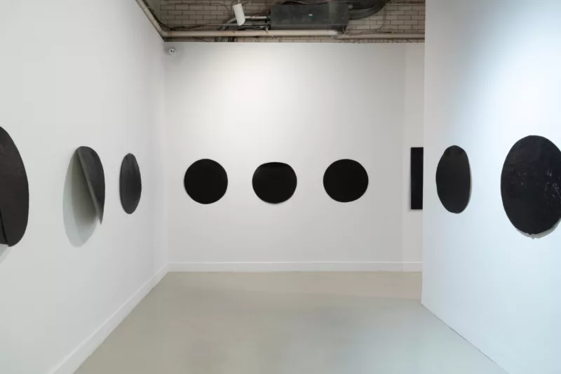 A gallery with a concrete floor and exposed ceiling pipes shows an installation of many circular black objects sitting on a white wall, one piece on the left wall seeming to pull away from its flat placement.