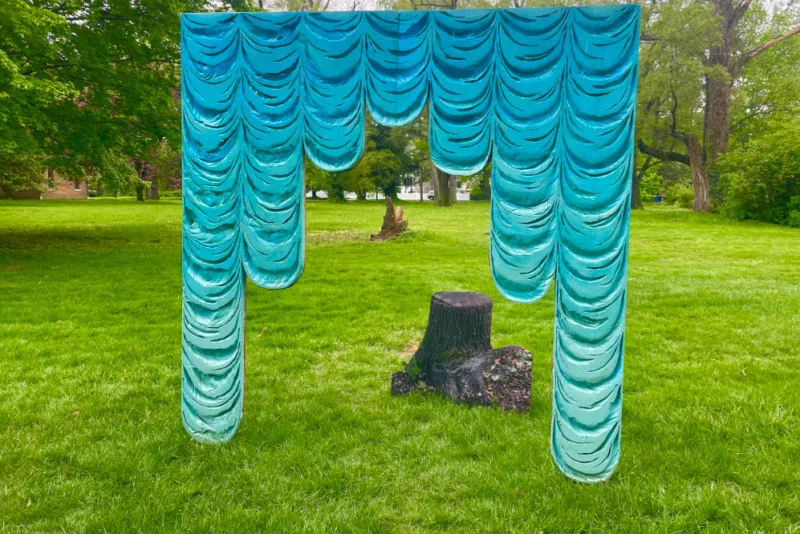 A setting in a grassy field with trees in the background shows a faux, painted theatrical curtain in shades of aqua that is jarring and clashes with the green of the natural setting. Through the raised center panels of the faux theatrical curtain are spied a darkened tree stump and in the way back, another tree stump, this with a jagged top from being split by lightening.