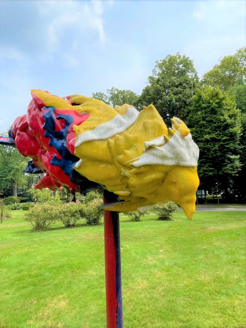 One of two colorful, drooping, bird-like objects that sit on top of tall poles situated close together in a grassy area surrounded by trees.