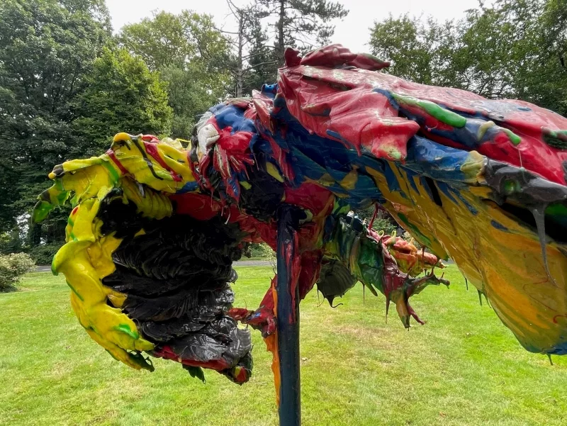 One of two colorful, drooping, bird-like objects that sit on top of tall poles situated close together in a grassy area surrounded by trees.