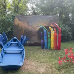 In a wooded area, a theatrical backdrop is set up with a tableau in front of it of a bright-colored, faux rainbow on the right and a bright blue rowboat and faux waves aiming at the picture as if ready to shoot the rapids in the river on the left. Fluttering red butterflies swirl in the foreground.